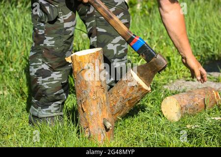 Holzfäller. Ein älterer Mann in Tarnhose hackt Holz mit einer alten Metallaxt, Beil mit einem Holzgriff in der Natur. Baumstümpfe von Pinien auf gr Stockfoto