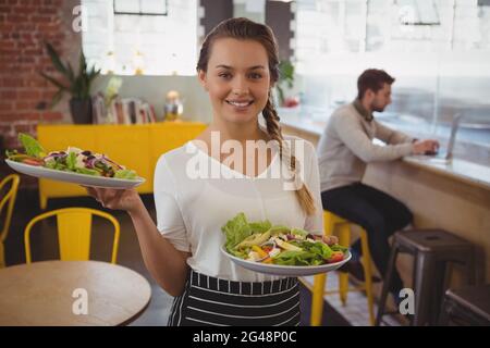 Porträt einer Kellnerin, die Teller mit Salat hält, während ein Geschäftsmann einen Laptop benutzt Stockfoto