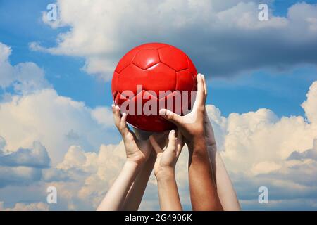 Eine Gruppe von Fußballfans, die roten Fußball in blau bewölktem Himmel halten. Die Hände von Kindern und Erwachsenen halten den Ball zusammen Stockfoto