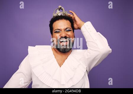 Porträt einer Drag Queen lächelt auf violettem Hintergrund. Bärtiger Mann mit Make-up und Krone auf dem Kopf. Stockfoto