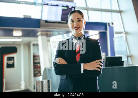 Porträt einer glücklichen jungen Frau, die am Flughafen arbeitet. Die Flugbegleiterin steht am Flugsteig und ihre Arme werden am internationalen Flughafen gekreuzt. Stockfoto