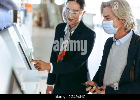 Eine Frau, die einen Gesichtsschutz trägt, hilft dem Geschäftsmann in einer Gesichtsmaske mit einem Check-in-Automaten am Flughafenterminal während einer Pandemie. Stockfoto