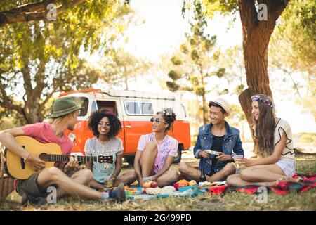 Lächelnde Freunde, die auf dem Feld sitzen und den Mann beim Gitarrenspiel betrachten Stockfoto