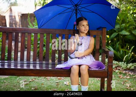 Mädchen, das mit blauem Regenschirm auf einer Holzbank sitzt Stockfoto