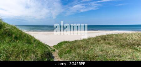 Ein Wanderweg führt durch hohe grasbewachsene Sanddünen zu einem abgeschiedenen und leeren weißen Sandstrand mit einem ruhigen türkisfarbenen Meer dahinter Stockfoto