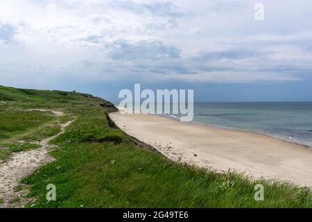 Der Wanderweg führt durch hohe grasbewachsene Sanddünen zu einem abgeschiedenen und leeren weißen Sandstrand mit einem ruhigen türkisfarbenen Meer dahinter Stockfoto