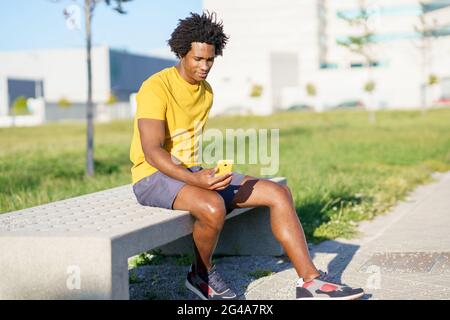 Schwarzer Mann, der sein Smartphone mit einer Übungs-App konsultiert, während er sich von seinem Training ausruhte. Stockfoto