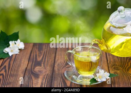 Grüner Tee mit Jasminblüten, die aus einer Glasküche in eine Tasse gießen Stockfoto