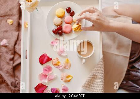 Romantisches Frühstück im Bett zum Valentinstag. Draufsicht auf die Hand der Frau, die Makronen hält. Duftender Kaffee, Rosenblüten, Dessertplatte und Beeren auf weißem Tablett Stockfoto