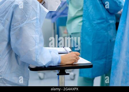 Eine Ärztin in blauer Uniform und einer medizinischen Maske macht während der Operation Notizen in Dokumenten. Stockfoto