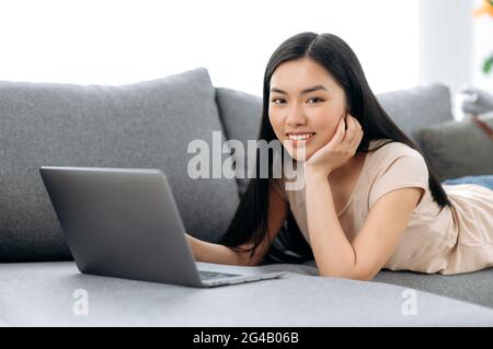 Charmante süße Brünette Mädchen von asiatischer Ethnie, liegt auf der Couch auf ihrem Bauch, nutzt einen Laptop, durchsucht soziale Netzwerke, Studien aus der Ferne, macht eine Bestellung nach Hause, sieht die Kamera, lächelt Positivität Stockfoto