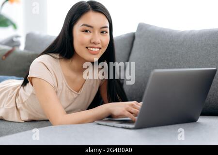 Hübsches, positives chinesisches oder japanisches Brünette-Mädchen liegt zu Hause auf der Couch auf dem Bauch, nutzt einen Laptop, durchstösst das Internet, chattet in sozialen Netzwerken, schaut die Kamera an, lächelt freundlich Stockfoto