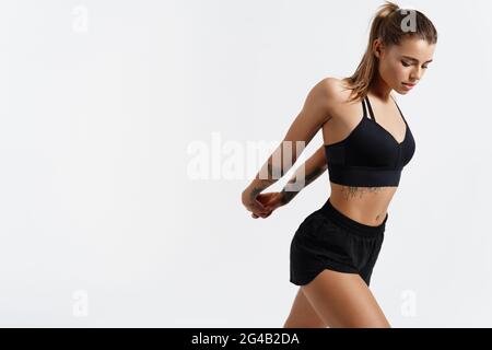 Schöne Fitness-Frau, weibliche Athletin, die sich die Hände nach hinten streckt, Aufwärmen für das Fitnesstraining, Training in Innenräumen, in schwarzer Sportkleidung, im Stehen Stockfoto