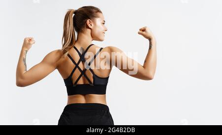 Sport und Frauen. Rückansicht einer starken Fitness-Athletin, einer Bodybuilderin, die Muskeln beugt, einen fitgen Körper, Bizeps und einen athletischen Rücken zeigt, lächelnd Stockfoto