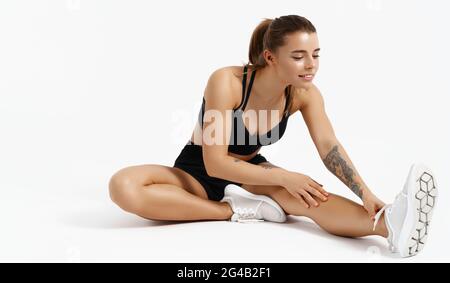 Bild einer jungen Fitness-Frau, die auf dem Boden sitzt und sich vor dem Joggen das Bein streckt. Der Läufer bereitet den Körper auf die Trainingseinheit vor und lächelt zufrieden Stockfoto