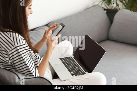Eine kurze Aufnahme einer Frau, die bequem zu Hause sitzt, mit einem Laptop und einem Smartphone. Mädchen beantworten Nachricht, chatten auf Handy, während Sie mit dem Computer arbeiten Stockfoto
