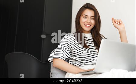 Bild von ehrgeizigen freiberuflichen Mädchen zu Hause sitzen mit Laptop und Blick auf die Kamera lächelnd. Frau, die im Wohnzimmer am Computer arbeitet Stockfoto