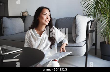 Ausruhen asiatische Frau lesen Magazin zu Hause, sitzen im Wohnzimmer am entspannenden Wochenende und Blick nach draußen Fenster, Freizeit im Haus, mit Stockfoto