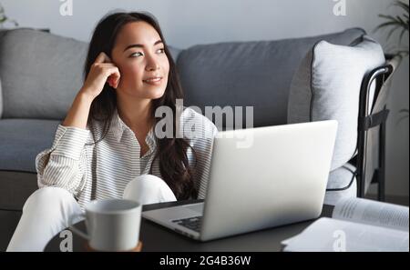 Asiatische Frau, die mit Computer arbeitet. Mädchen sitzt in der Nähe des Laptops zu Hause, blickt aus dem Fenster und lächelt entspannt, am Wochenende ausruhen, Videos anschauen Stockfoto