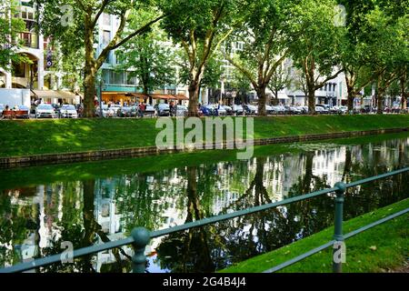 Die berühmte Einkaufsmeile Königsallee: Blick über den grünen Stadtkanal 'Kö-Graben' von der 'ruhigen Seite' auf die 'belebte Seite' mit vielen Geschäften. Stockfoto