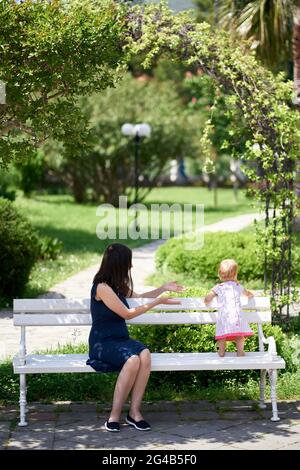 Das kleine Mädchen steht auf einer Parkbank neben ihrer sitzenden Mutter Stockfoto