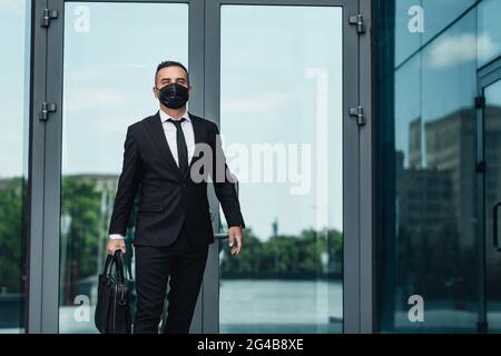Serious Geschäftsmann in schützende Gesichtsmaske hält Aktentasche und verlassen moderne Bürogebäude Stockfoto