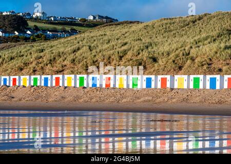 Weiße Strandhütten, mit farbigen Türen, stehen vor Dünen mit Reflexen im feuchten Strandsand am Woolacombe Beach, Devon Stockfoto