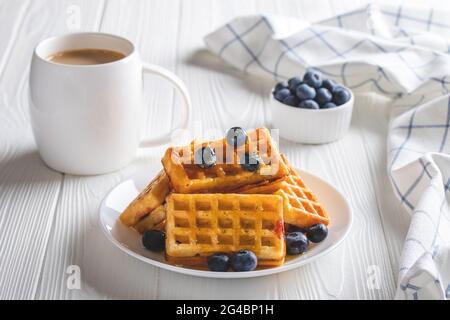 Belgische Waffeln mit Heidelbeeren und Kaffee auf einem Holztisch. Warmes Morgengetränk und Beeren, Frühstückskonzept. Latte in einer weißen Tasse und Nahaufnahme Stockfoto