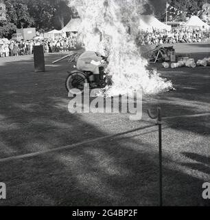 1960er Jahre, historisch, draußen auf einem Feld, bei einer County Show oder Fete, Zuschauer beobachten ein Motorrad-Display, mit einem der Display-Team reiten durch Feuer, ein Hochrisiko, dramatische waghalsige Stunt, England, Großbritannien. Motorrad-akrobatische Darsteller bei Karnevals und Sonderveranstaltungen waren in dieser Zeit beliebt, mit den Royal Signals, den "Weißen Helmen", einem Armee-Display-Team, das bekannteste. Sie hatten die erste aufgezeichnete Stunt-Ausstellung 1928 durchgeführt, als die Dispatch-Fahrer, die ein wichtiger Teil der Kommunikation der Armee waren, hochqualifizierte Motorradfahrer sein mussten. Stockfoto