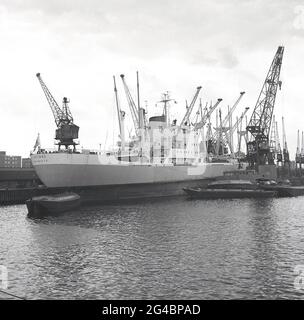 Das historische griechische Frachtschiff IKTINOS aus den 1960er Jahren vertäute an den Tilbury Docks an der Themse in Tilbury Essex, England, einem Teil des größeren Hafens von London. Daneben der PLA-Schwebekran London Leviathan, ein selbstfahrender Krankahn, der 1965 in Amsterdam für die Port of London Authority gebaut wurde. Das Frachtschiff IKTINOS wurde im Juli 1969 von Doxford & Sunderland Shipbuilding & Engineering Co., Ltd., Sunderland, UK, für Lineas Interoceanicas S.A. unter griechischer Flagge gebaut. Stockfoto