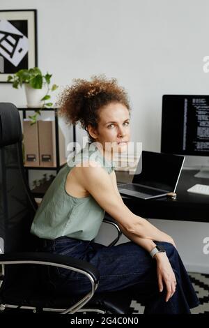 Porträt einer jungen Frau, die auf einem Stuhl sitzt und die Kamera anschaut, während sie an ihrem Arbeitsplatz im Büro arbeitet Stockfoto