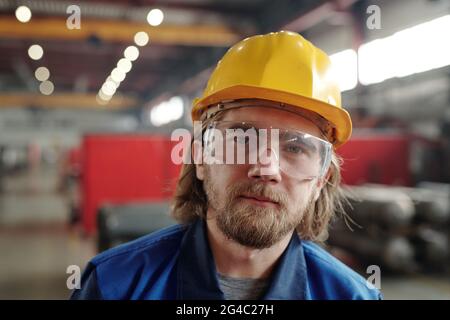 Porträt eines ernsthaften jungen bärtigen Fabrikmanns in gelbem Hardhut und einer Schutzbrille, die im Hintergrund gegen Gasflaschen posiert Stockfoto
