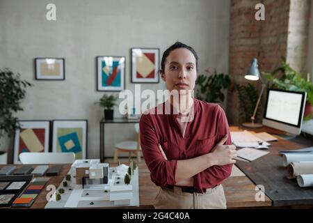 Porträt einer seriöse hispanischen Architektin, die mit gekreuzten Armen gegen einen Holztisch mit Mustern und einem 3D-Modell des Bürogebäudes steht Stockfoto