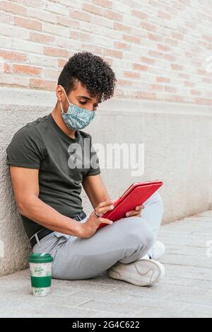Vertikales Porträt eines jungen lateinamerikanischen jungen Mannes mit einer Gesichtsmaske. Sitzend auf dem Boden und mit einem Tablet Stockfoto