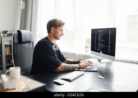Junger männlicher Programmierer, der Programmcode schreibt, sitzt am Arbeitsplatz mit Monitor im Büro Stockfoto