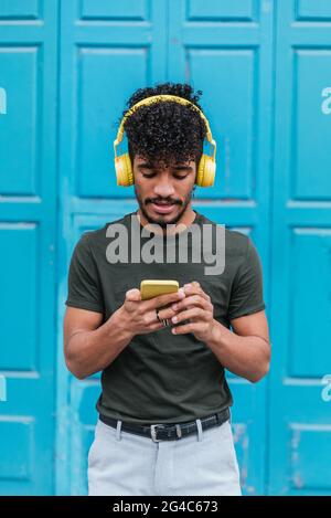 Vertikales Porträt eines jungen lateinamerikanischen Mannes, der auf der Straße ein Telefon mit gelben Kopfhörern und blauem Türhintergrund benutzt Stockfoto