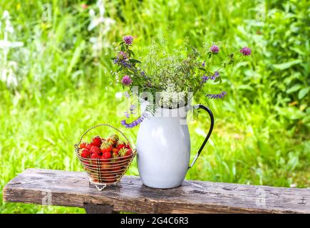 Erdbeeren in einem Korb und Blumenstrauß in einem Metallkrug auf einer Holzbank Stockfoto