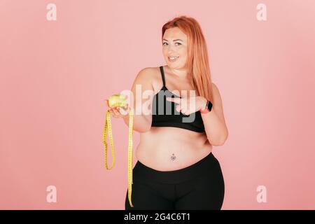 Die schöne glückliche Plus-Size-Frau trägt ein sportliches Top und Leggings und hält ein Apfel- und Zentimeterband auf pinkem Studiohintergrund. Stockfoto