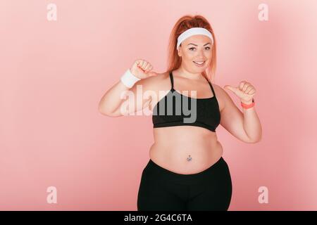 Plump glückliche Frau trägt sportliche Outfit lächelnd und zeigt Daumen nach oben, während sie vor rosa Studio Hintergrund stehen. Stockfoto