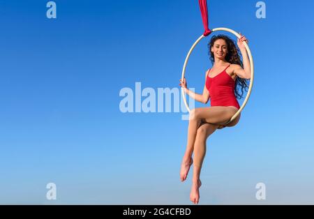 acrobat athletische, junge anmutige Turnerin Porträt auf Luftring im Freien auf Himmel Hintergrund. Flexible Frau in rotem Anzug führt Zirkus Künstler tanzen Stockfoto