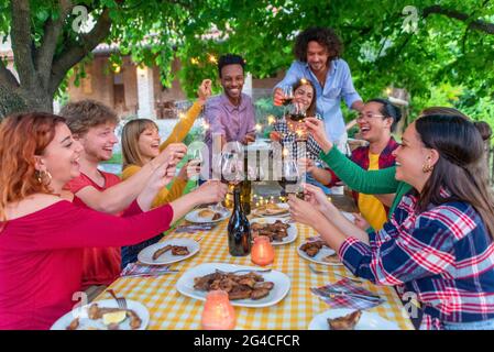 Fröhliche Gruppe von Menschen feiern Sommerferien beim Barbecue-Abendessen Toasting Wein und halten Wunderkerzen im Freien. Diverse Freunde sitzen auf Picknick Stockfoto