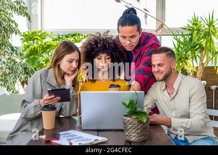 Eine Gruppe junger Erwachsener diskutiert im Coworking Space Indoor über ein ernsthaftes Startup-Projekt. Diverse Studenten arbeiten bei der Arbeit an Laptop und Tablet zusammen Stockfoto