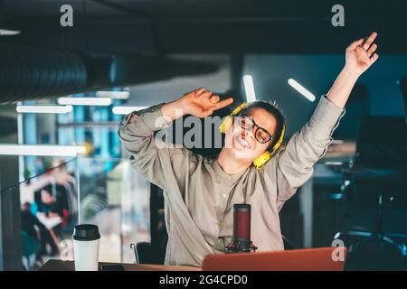 Attraktive junge Frau in den Dreißigern, die eine Brille und gelbe Kopfhörer trägt. Fröhliches Singen und Tanzen an ihrem Arbeitsplatz mit erhobenen Händen, Porträt Stockfoto