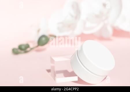 Handcreme, Feuchtigkeitscreme, Lotion im weißen Glas auf rosa Blumenhintergrund, Vorderansicht. Anti-Aging-Kosmetik-Produktvorlage. Einrichtung mit Acrylblock und Orchideen Stockfoto