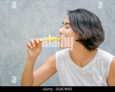 Frau, die Eis isst. Glücklich schöne asiatische Frau trägt lässige weiße ärmellose Hemd mit gelben Popsicle, im Freien. Lächelnde Frau, die mich genießt Stockfoto