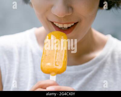 Nahaufnahme gelbe Popsicle in schönen glücklich asiatischen Frau die Hand trägt lässige weiße ärmellose Hemd, im Freien. Frau, die Eis isst. Lächelnd, weiblich Stockfoto