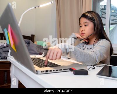 Asiatische Mädchen in Kopfhörer sitzen am Schreibtisch Studie online auf Laptop. Kinder tragen Headset-Handschrift beim Lernen im Notebook mit Hilfe von Internet-Lektionen über Quarantäne. G Stockfoto