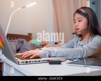 Asiatische Mädchen in Kopfhörer sitzen am Schreibtisch Studie online auf Laptop. Kinder tragen Headset Tastatur eingeben Notebook lernen mit Internet-Unterricht in Quarantäne. Stockfoto