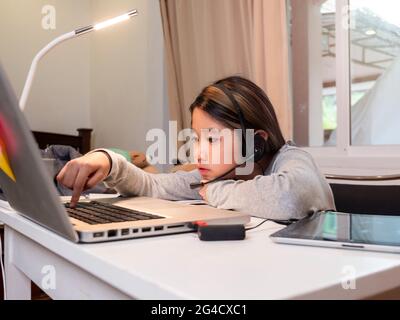 Asiatische Mädchen in Kopfhörer sitzen am Schreibtisch Studie online auf Laptop. Kinder tragen Headset-Handschrift beim Lernen im Notebook mit Hilfe von Internet-Lektionen über Quarantäne. G Stockfoto