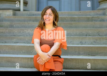 Trendige Frau mittleren Alters im farbenfrohen orangefarbenen Sommer-Outfit, die auf der Außentreppe wartet und mit einem nachdenklichen Lächeln mit Copyspace zur Seite schaut Stockfoto